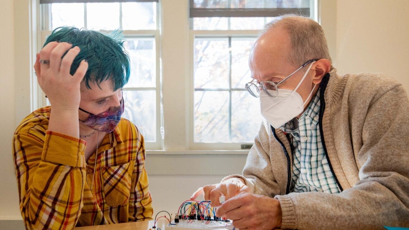 An older man wearing a mask explains an electronic circuit board to a younger person with teal hair and a plaid shirt at a table by a window.