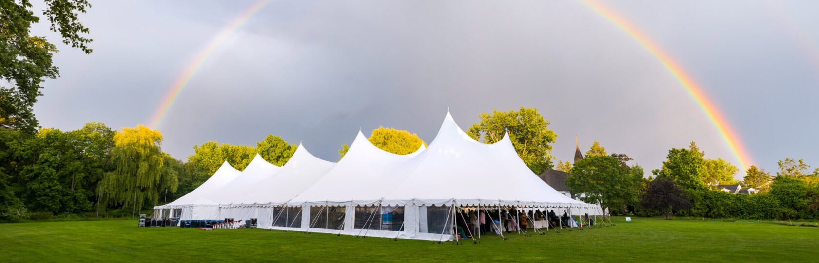 A large white event tent is set up on a green lawn with a double rainbow arching across the cloudy sky overhead.