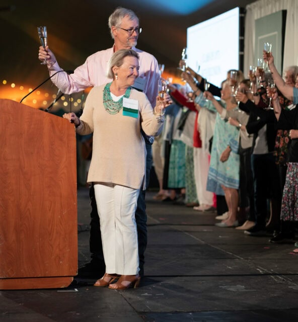 A group of people stand on a stage holding raised glasses in a toast, with a man and woman at a wooden podium leading the group. The stage is decorated with string lights and a large screen in the background.