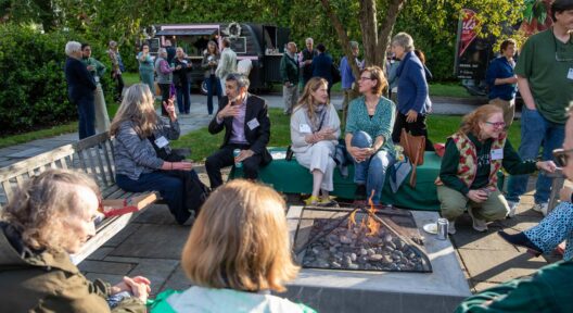 A group of people sitting around a fire pit in an outdoor setting, talking and engaging with one another. Trees, a food truck, and other attendees are visible in the background.