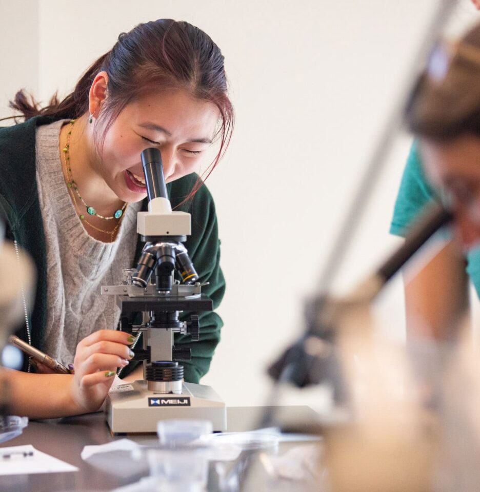 A young woman smiles while looking through a microscope in a laboratory setting, surrounded by other students.