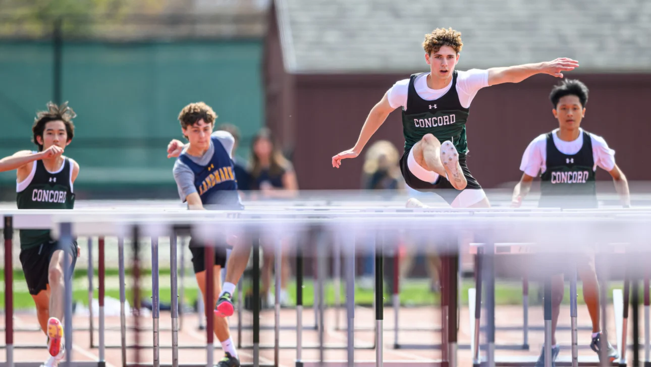 Four athletes compete in a hurdling race on a track. One athlete is in mid-air, clearing a hurdle, while the others follow closely behind.