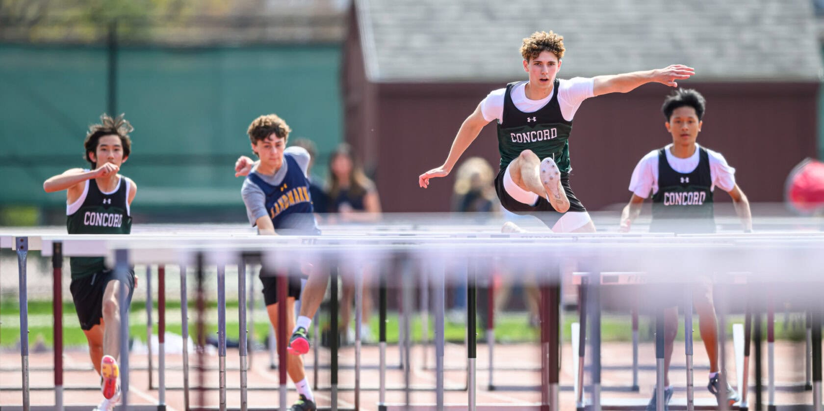 Four athletes compete in a hurdling race on a track. One athlete is in mid-air, clearing a hurdle, while the others follow closely behind.