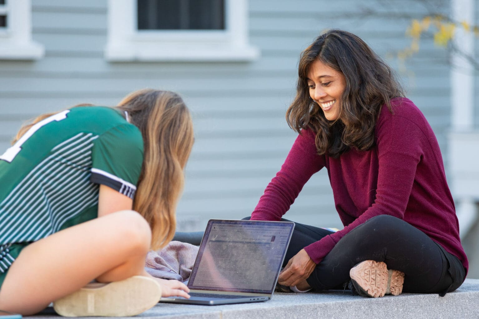 Two people sitting outdoors, one using a laptop. The person on the left is wearing a green striped shirt and facing away, while the person on the right is smiling and wearing a maroon sweater.