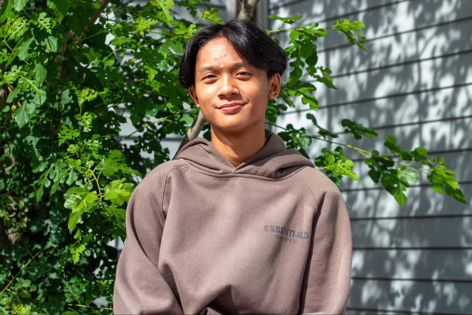 A person wearing a brown hooded sweatshirt stands in front of a leafy green background and a gray wall.