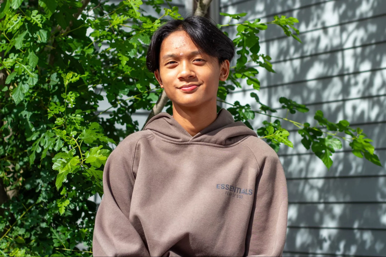 A person wearing a brown hooded sweatshirt stands in front of a leafy green background and a gray wall.