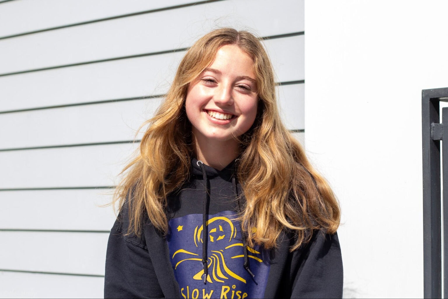 A young person with long, wavy blonde hair smiles while wearing a black sweatshirt with a graphic design, standing in front of a striped wall.