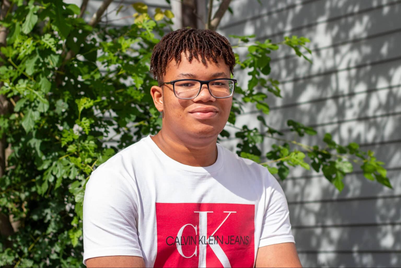 Young person with short braided hair and glasses wearing a white Calvin Klein Jeans shirt, standing outside in front of greenery and a building.