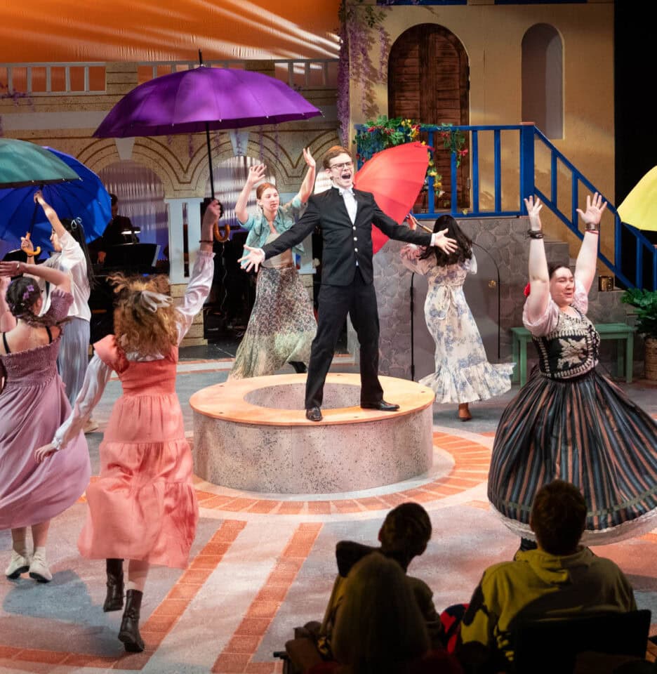 A theatrical performance with actors in colorful costumes dancing around a man in a suit standing on a circular platform. Some actors are holding vibrant umbrellas. An audience watches from the foreground.