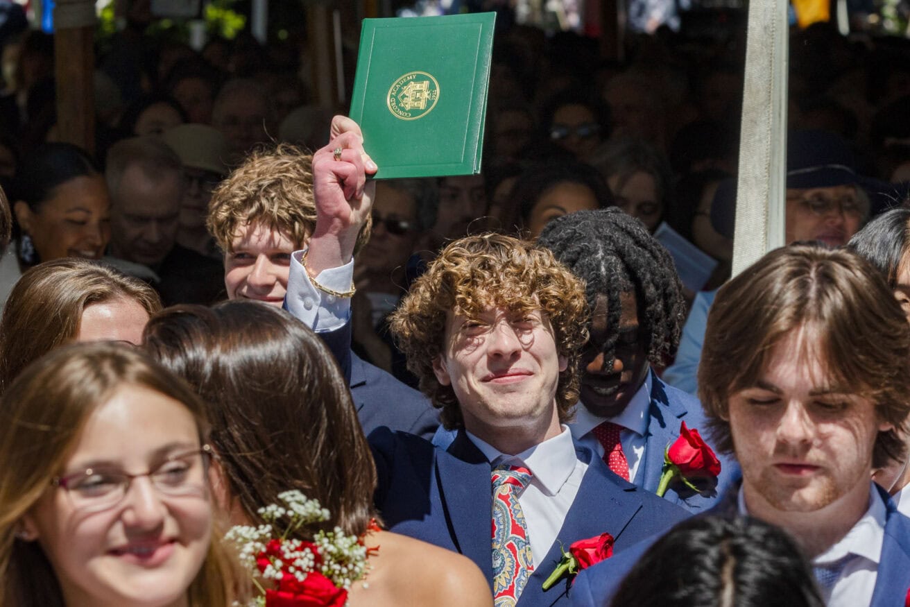 A young man with curly hair, wearing a suit and tie, holds up a green diploma cover while surrounded by other graduates. People are smiling and celebrating in the crowded scene.