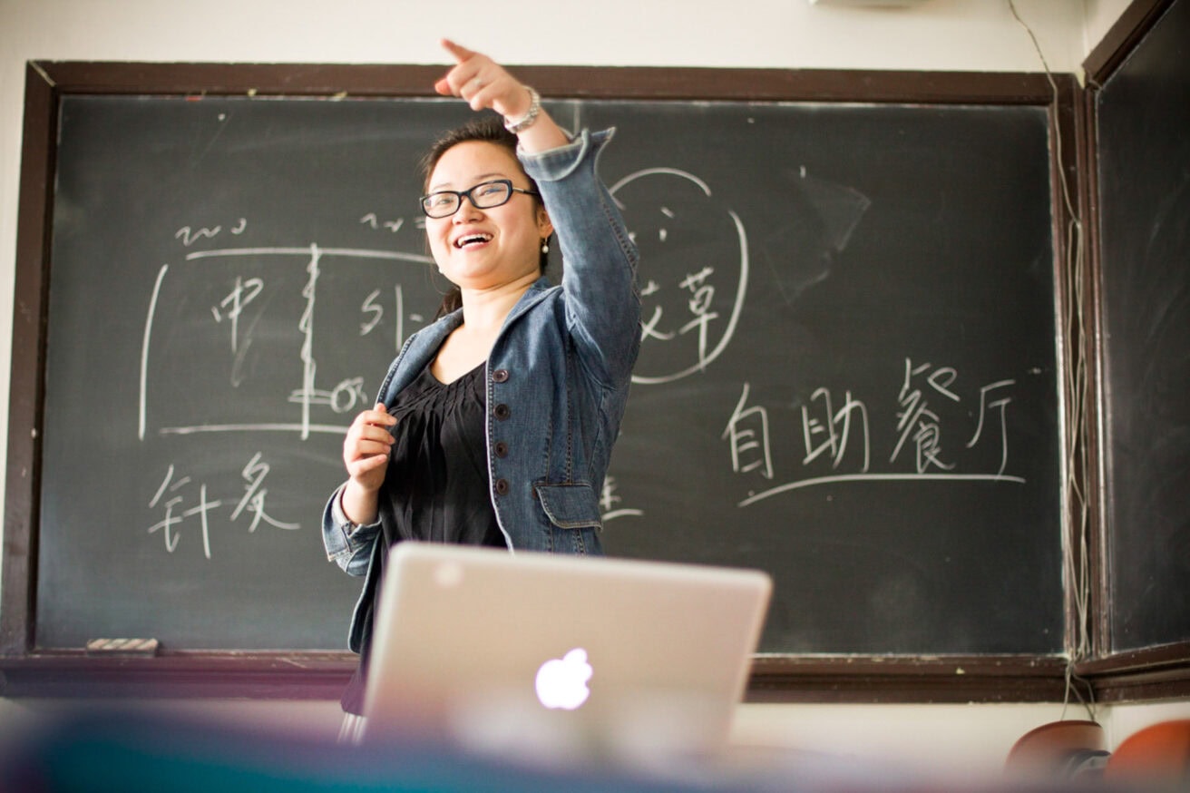 A smiling person in a denim jacket stands in front of a chalkboard with writing on it, pointing towards something off-camera. An open laptop is in the foreground.