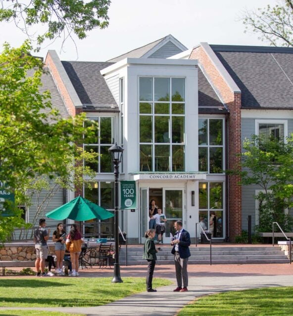 A group of people stand and sit near the entrance of a building labeled "Concord Academy" surrounded by greenery on a sunny day.