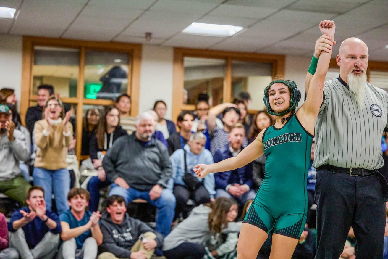 A wrestling athlete in a green singlet is declared the winner by a referee lifting their arm, with cheering spectators in the background.