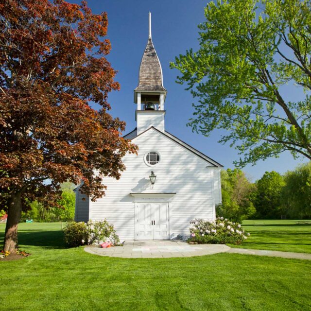 A small white chapel with a steeple stands amid lush green trees and a well-maintained lawn on a sunny day.