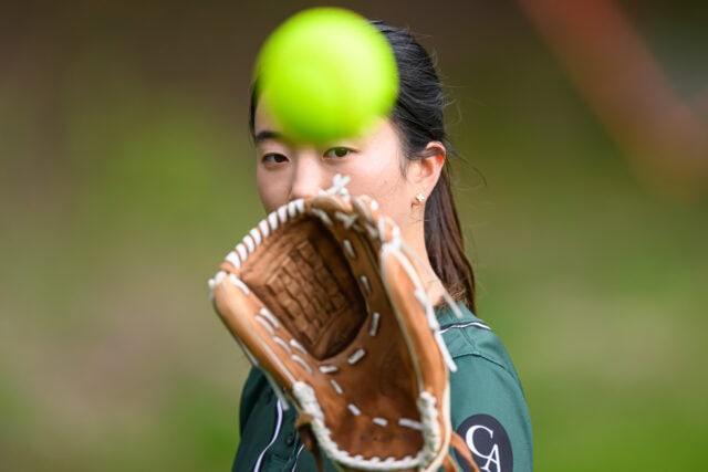 A person wearing a green shirt catches a yellow softball with a brown glove. The focus is on the ball and glove, with the person's face partially visible in the background.