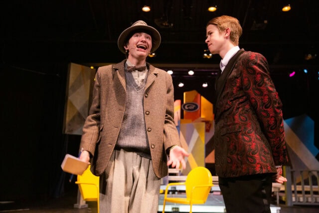 Two actors in period costumes perform on stage under dramatic lighting, with one holding a book and gesturing animatedly while the other listens attentively.