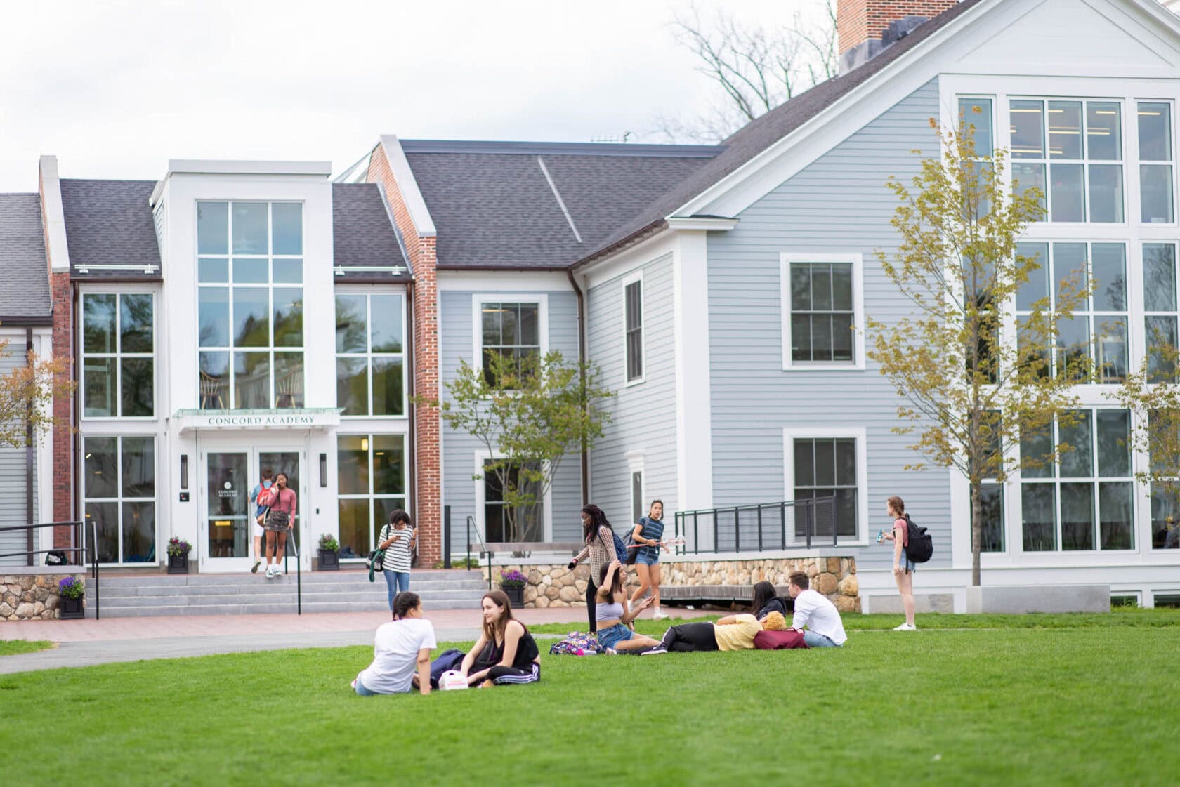 A group of students sits on the grassy lawn, with others walking nearby in front of a modern, multi-story building with large windows.