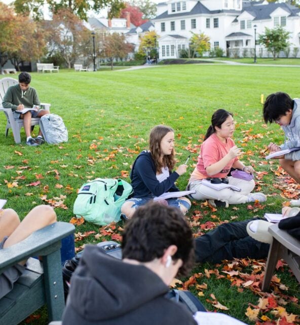 A group of students sit on a lawn, engaged in studying and writing. Some are seated on chairs while others sit on the ground. Leaves are scattered around, indicating a fall setting.