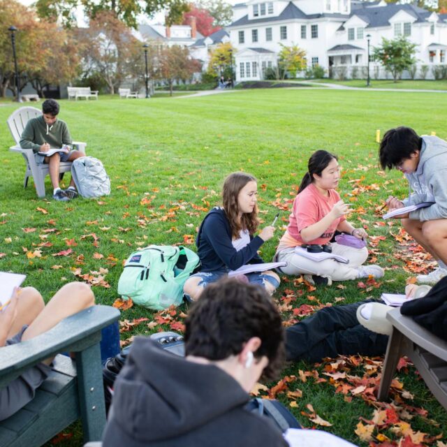 A group of students sit on a lawn, engaged in studying and writing. Some are seated on chairs while others sit on the ground. Leaves are scattered around, indicating a fall setting.