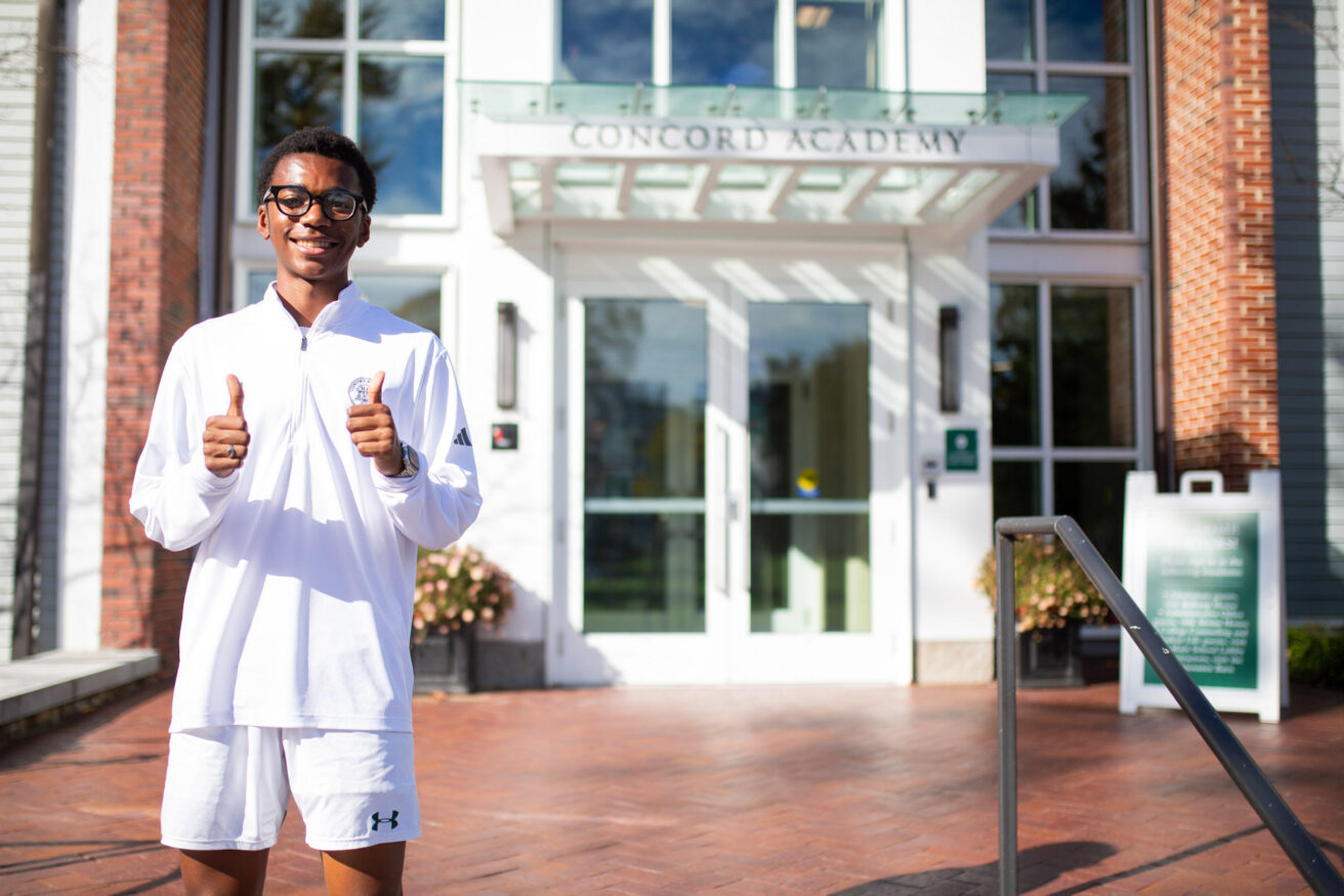 A person stands with thumbs up in front of a building with a sign reading "Concord Academy." The person is wearing glasses and a white athletic outfit.