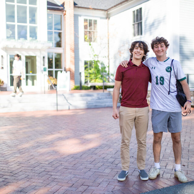 Two young men are posing and smiling in front of a school building. One wears a maroon shirt and khaki pants, while the other is in a white soccer jersey with the number 19.