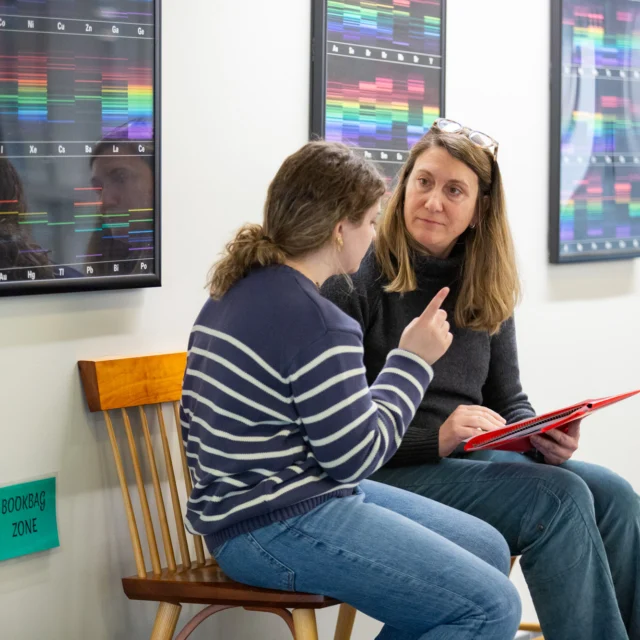 Two individuals are seated on wooden chairs in a hallway, engaged in conversation. One person is holding a tablet, and colorful charts hang on the wall behind them.