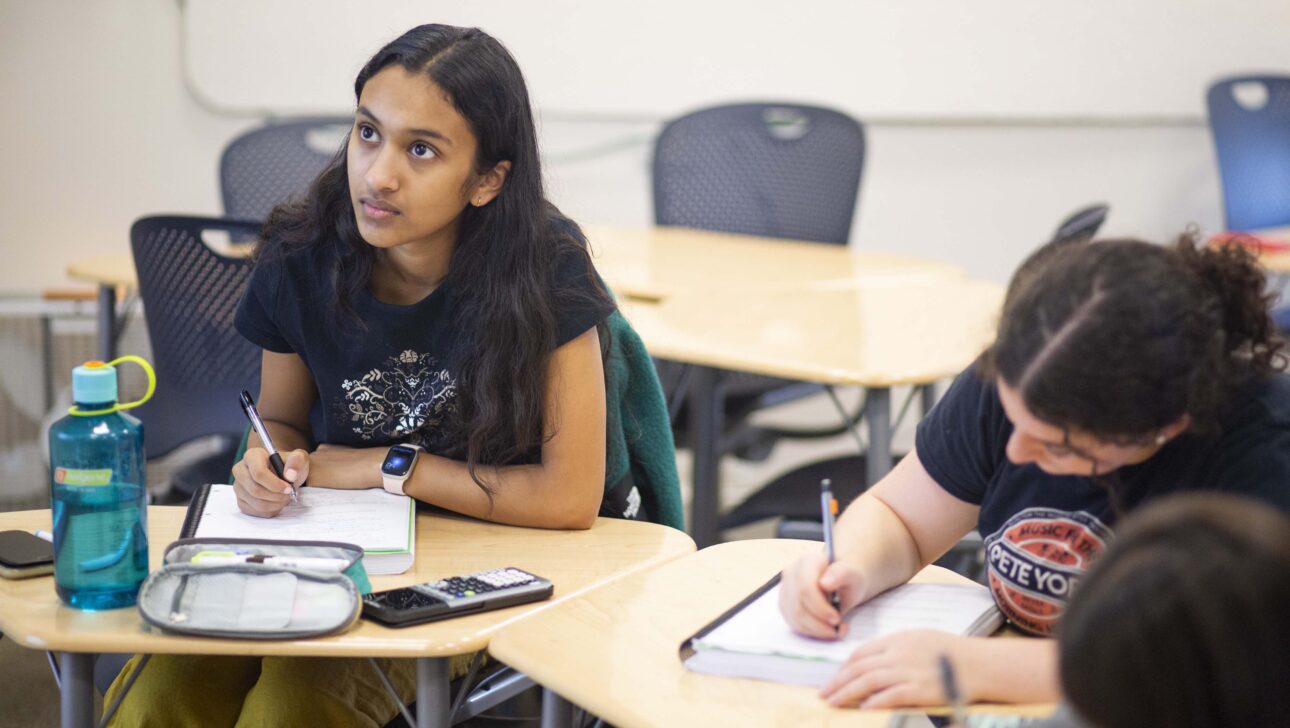 Students sit at desks in a classroom. Two are bent intently over their work, and another, a girl, looks up intently.