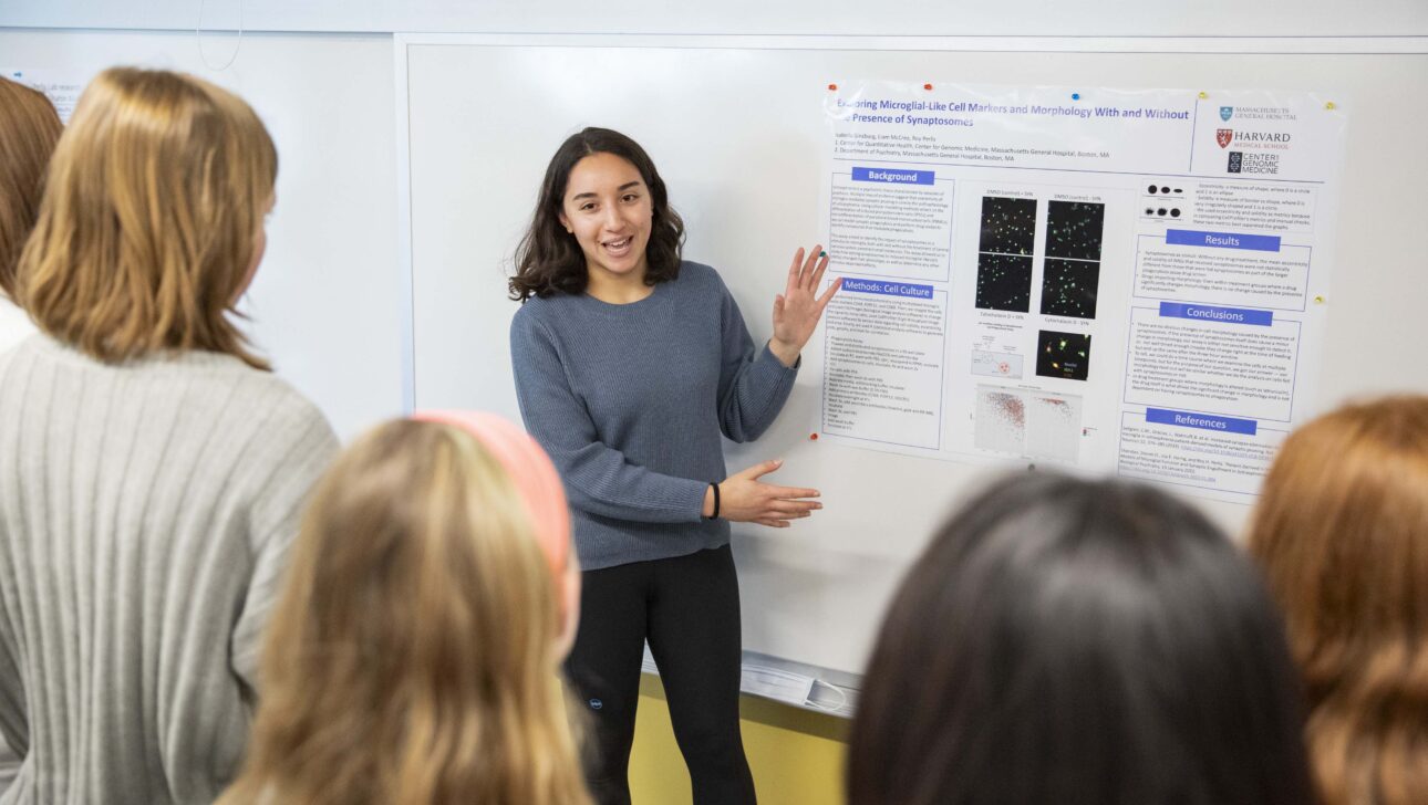 A student gestures to a poster hanging behind her, talking with a small group about her science internship.