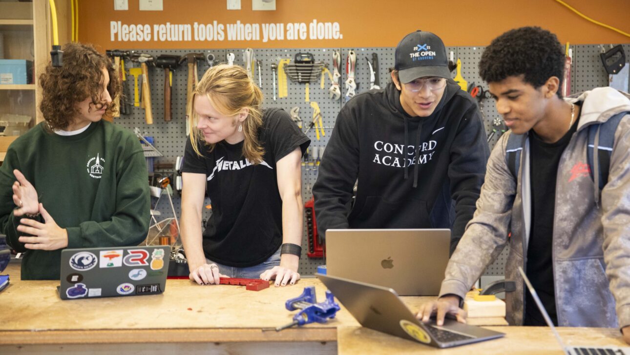 Four students lean over a table, paired off in conversation over laptops; behind them is a wall of tools.