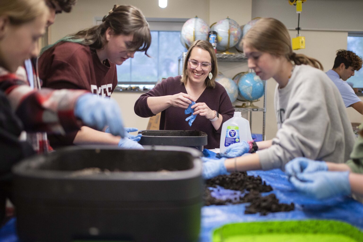 A teacher smiles while putting on a glove; her students are doing a vermiculture lab with live worms.