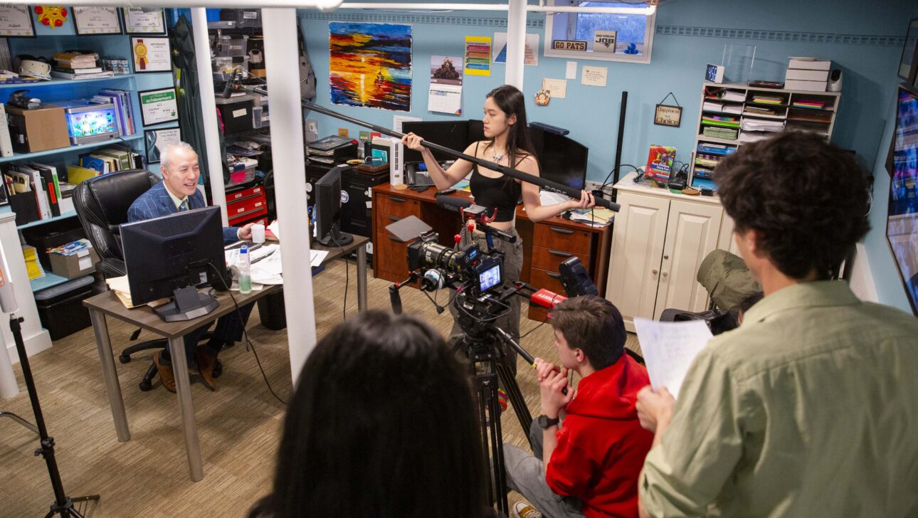 A female student holds a boom microphone while a male student uses a videocamera to film an actor in an office, while other crew members look on.