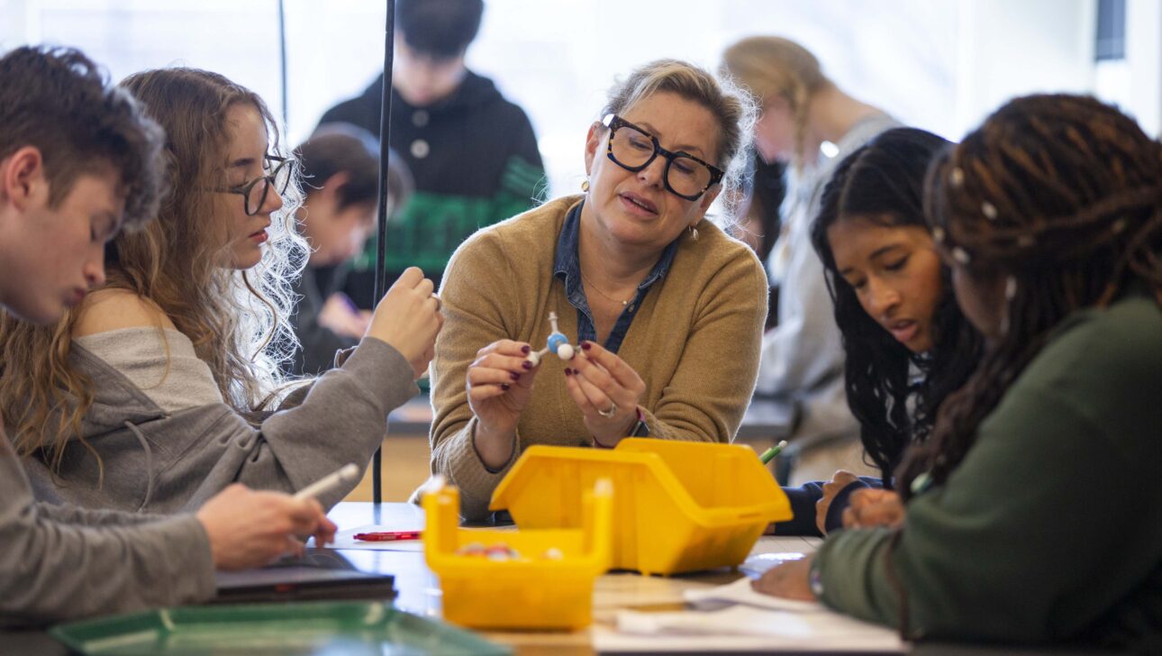 A teacher holds a model of a chemical bond over a yellow bin, examining it while students around the table look at similar pieces.