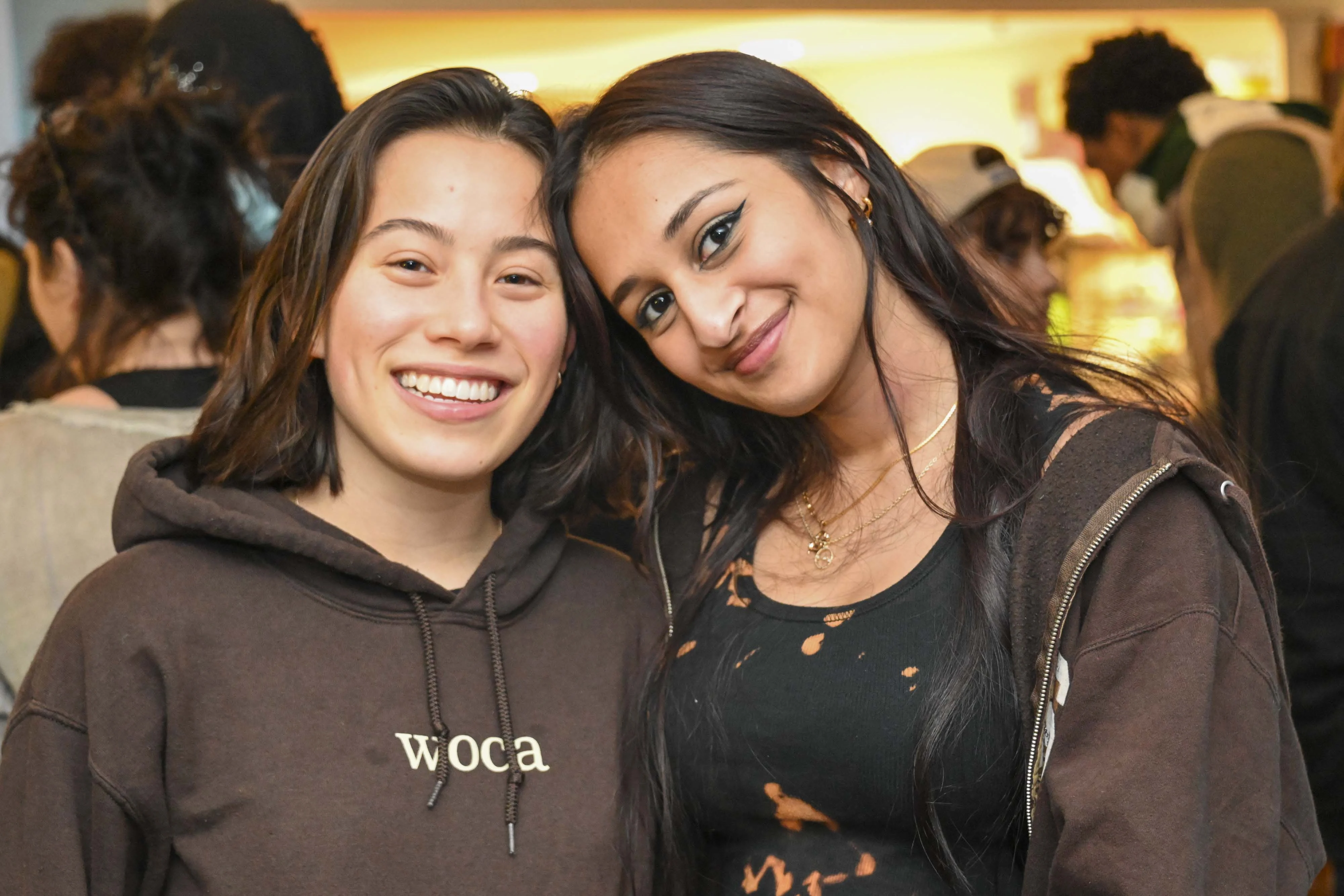 Two girls smile for the camera at a social event; one wears a sweatshirt that says WoCA, which stands for Women of Color Alliance.