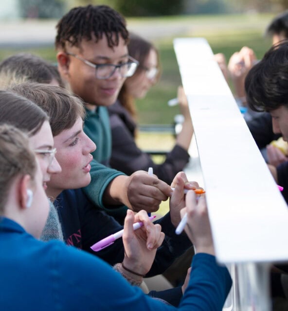 A group of people sit at a long table outside, each holding markers, engaged in an activity. They are attentively focused on the task at hand.