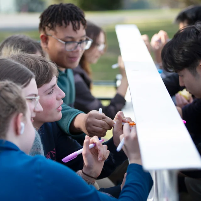 A group of people sit at a long table outside, each holding markers, engaged in an activity. They are attentively focused on the task at hand.