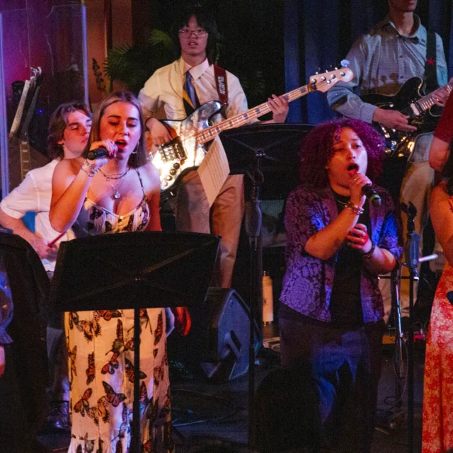A group of musicians performs on stage. Four vocalists sing into microphones at the front, while instrumentalists, including a bassist and a guitarist, play in the background.