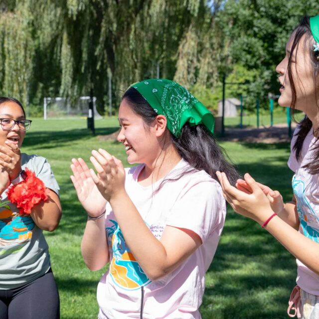 Three people wearing green bandanas and colorful shirts are standing outdoors on a sunny day, clapping and smiling. Trees and grass are visible in the background.