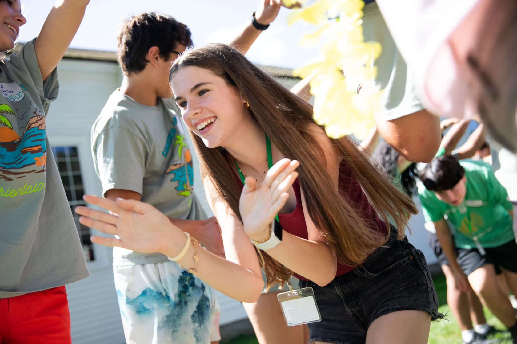 A young woman with long hair and a name tag participates in an outdoor group activity, smiling and clapping while others in casual attire engage in the background.