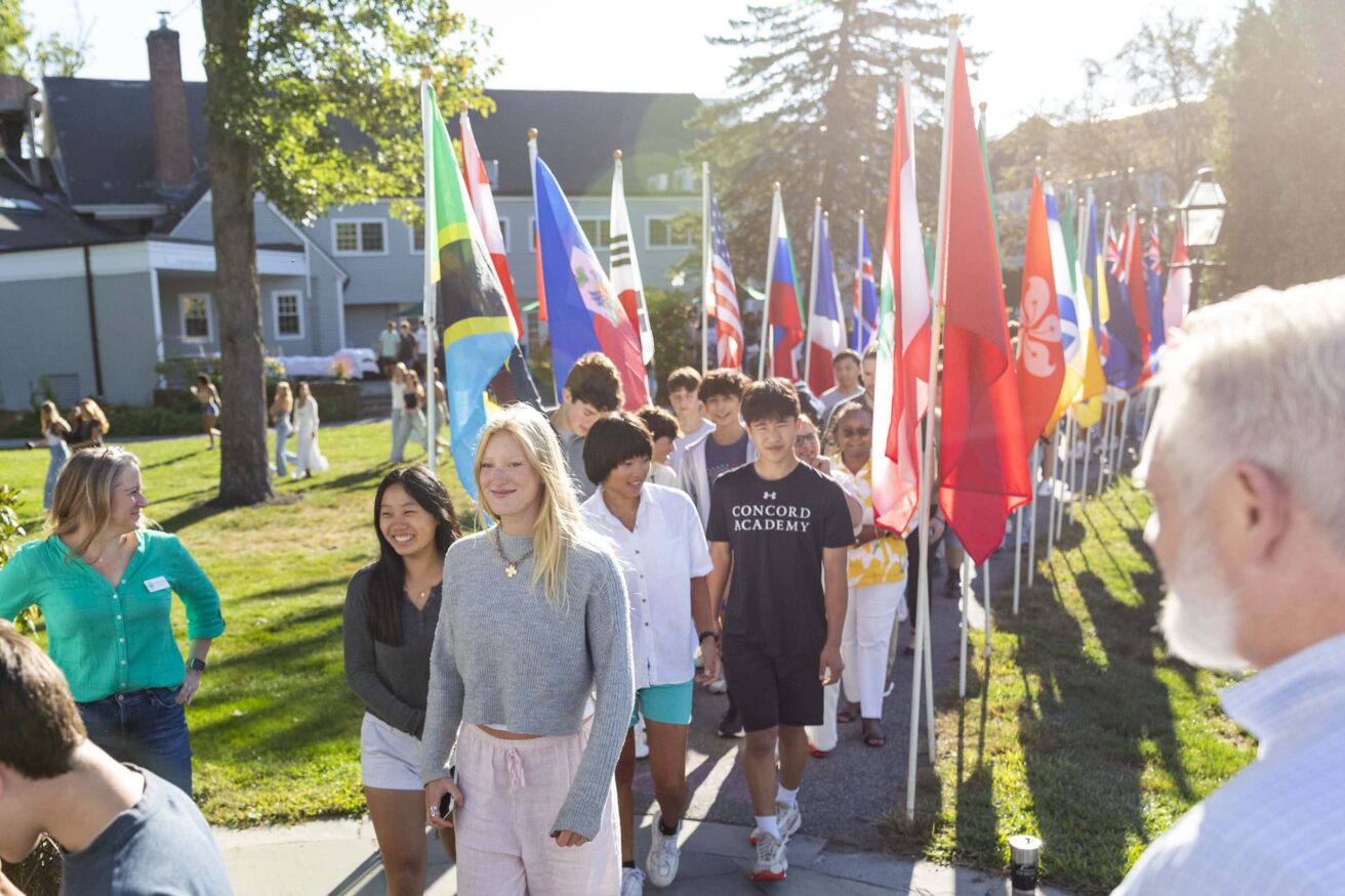 A group of students walking outdoors, passing by a row of international flags. Green lawn and buildings are in the background.