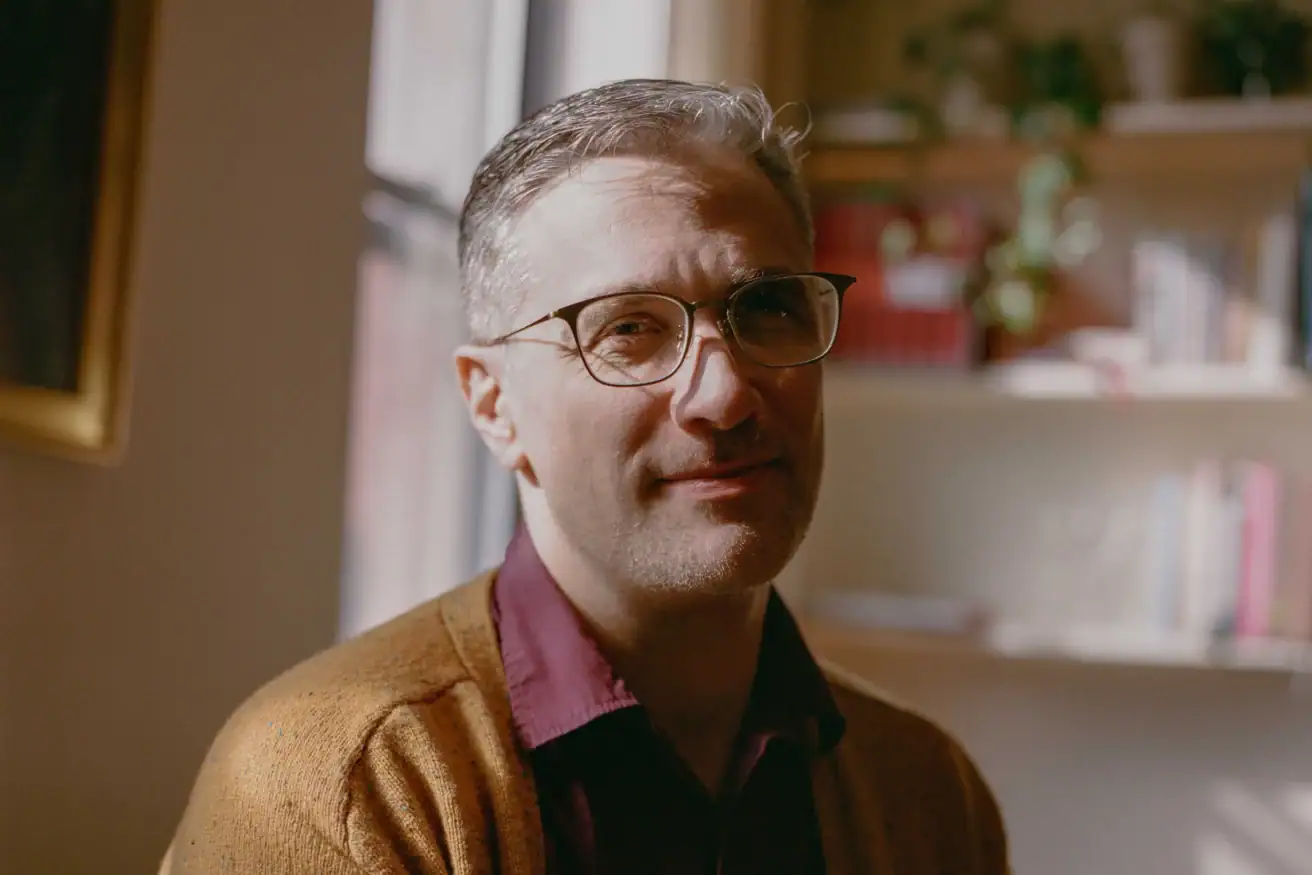 A white man in his 30s with graying hair and glasses, wearing a red collared shirt and orange sweater, sits half in shadow in an office, in front of a white bookcase