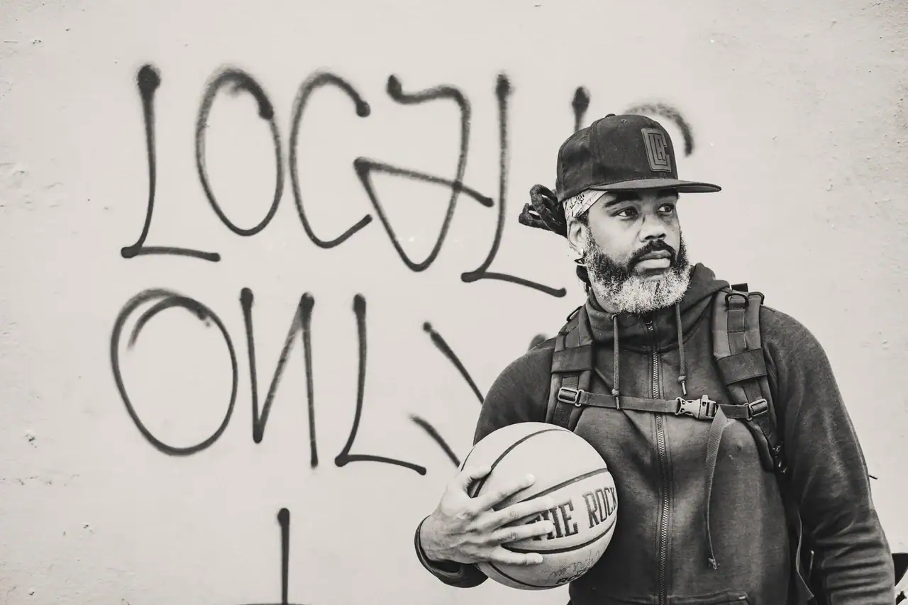 In black and white, a Black man wearing a hoodie and ball cap, holding a basketball and standing in front of a wall with graffiti, looks pensively off to the side