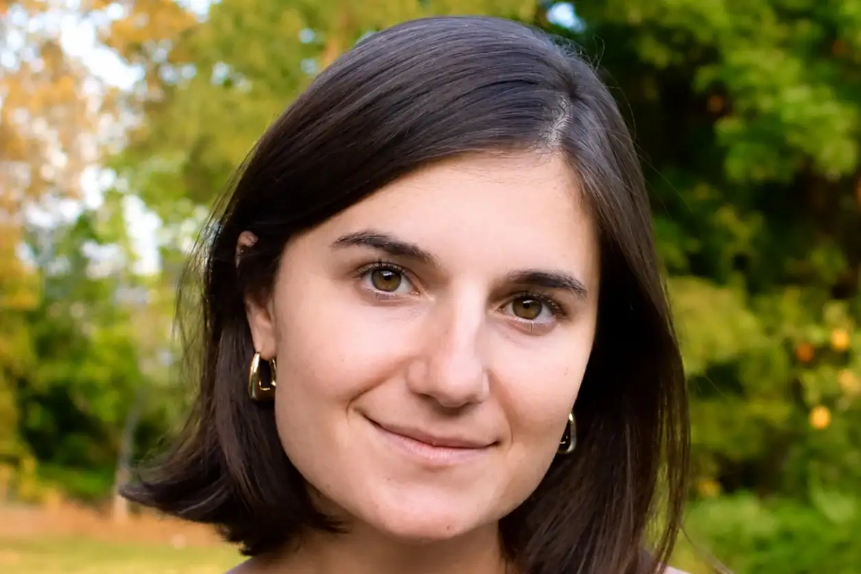 A young white woman with dark, shoulder-length hair and small hoop earrings, smiling in front of a backdrop of trees